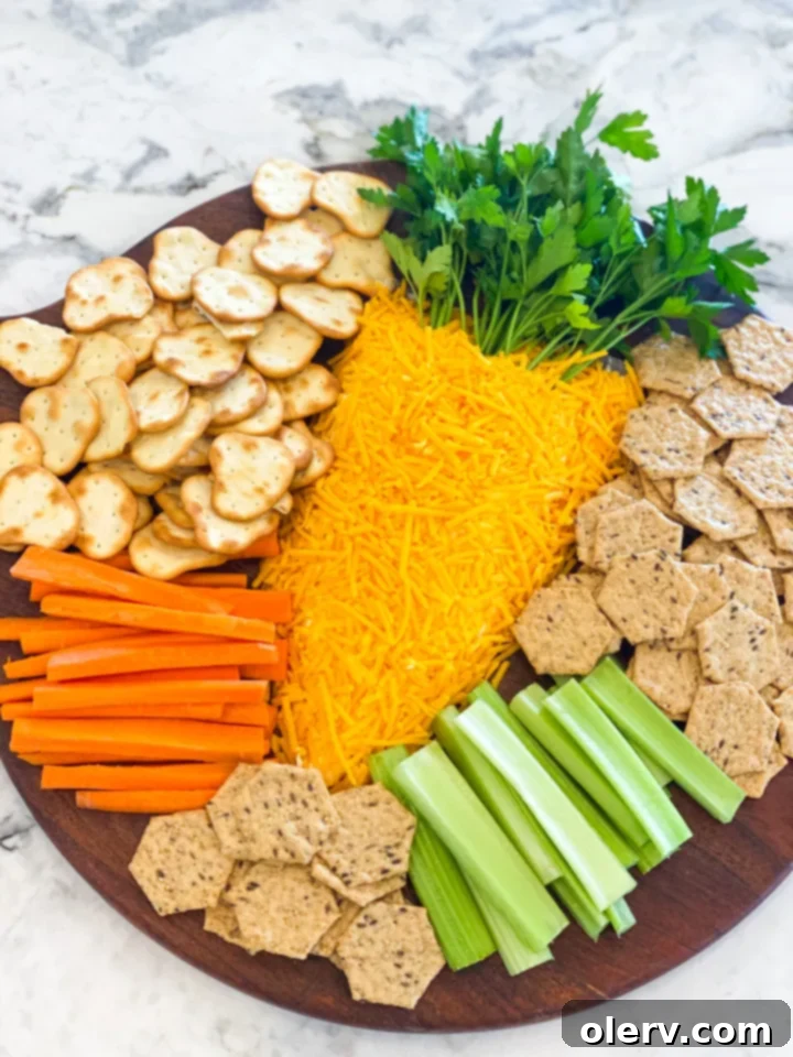 A festive Easter Carrot Cheese Ball shaped like a carrot, covered in shredded cheddar cheese with parsley for the green top, served on a white platter with various crackers and vegetables.