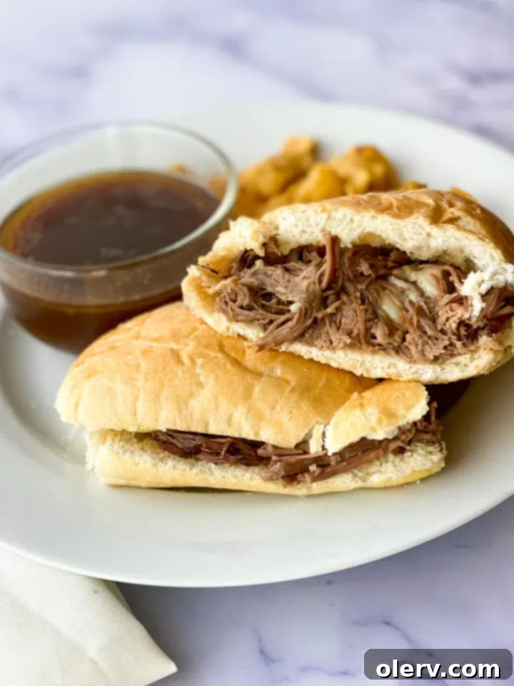 Ultimate Slow Cooker French Dip Sandwiches 7 Two beautifully prepared Slow Cooker French Dip sandwiches on a rustic plate, accompanied by a small bowl of rich au jus for dipping.